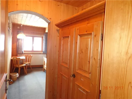 Ein heller Flur mit Holzwänden und einem Torbogen. Im Hintergrund sind ein Tisch und Stühle sowie ein Fenster zu sehen.