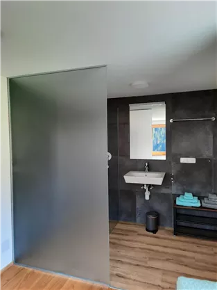 A modern bathroom with a semi-transparent shower wall and a sink. The floor is made of wood, and there is a stool with towels.