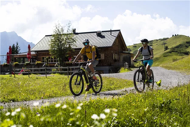 Two cyclists enjoy a tour in the mountains. In the background, a cozy wooden house and green meadows can be seen.