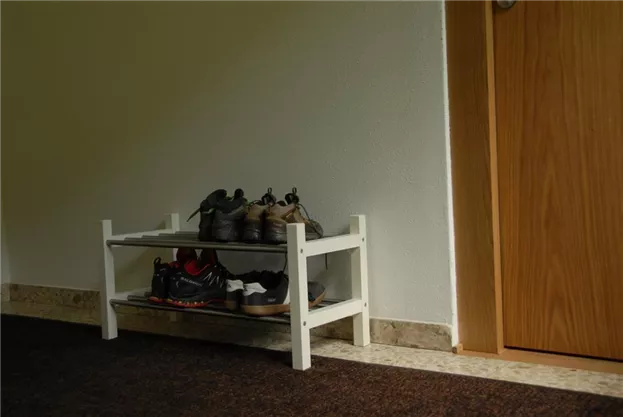 A shoe rack with several pairs of shoes is located in an entrance area. The wall is light, and the door is made of wood.