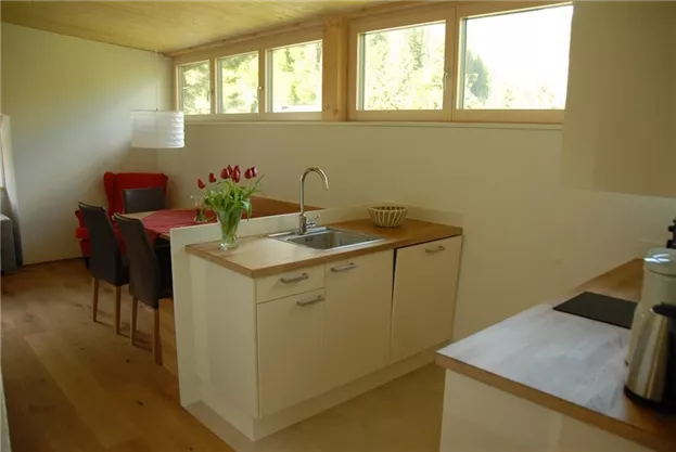 A modern kitchen with bright wood surfaces and large windows. A dining table with red chairs and a vase with fresh tulips are visible.