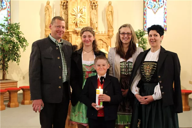 Eine Familie steht in einer Kirche, fröhlich zusammen mit traditioneller Kleidung. Ein Junge hält eine Kerze in der Hand, während die Erwachsenen lächeln.