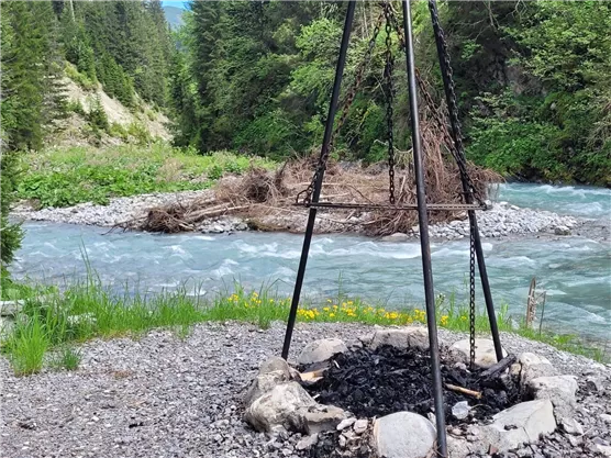 A campfire area with a firebench is next to a clear river. In the background, green trees and bushes can be seen.