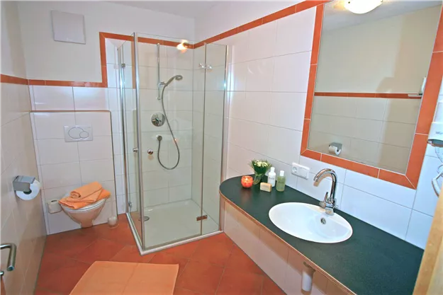A modern bathroom with a glass shower and a sink. The walls are light tiled and there are decorative elements on the shelf.