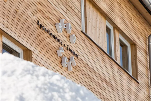 Ein Holzhaus mit einem Schild, das „Apartments & Zimmer“ zeigt. Der Schnee liegt im Vordergrund und verleiht der Szene eine winterliche Atmosphäre.
