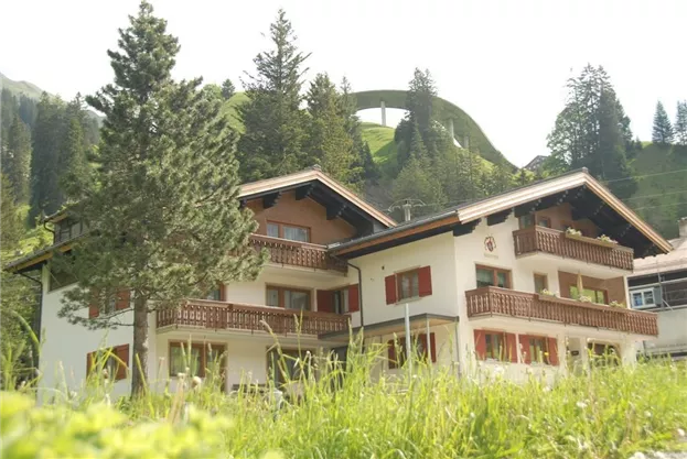 A charming mountain house with several balconies, surrounded by green meadows and tall trees. Gentle hills can be seen in the background.