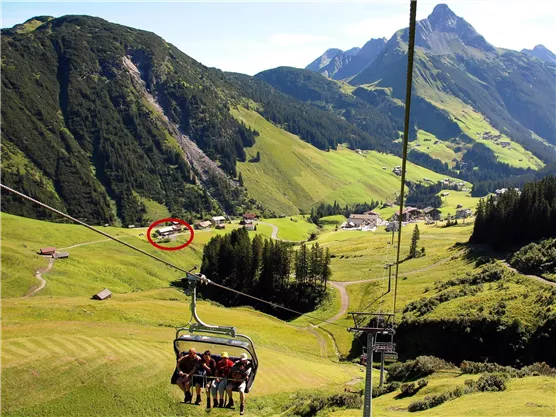 A cable car travels over green meadows and mountains. In the background, impressive mountain peaks and small huts can be seen.