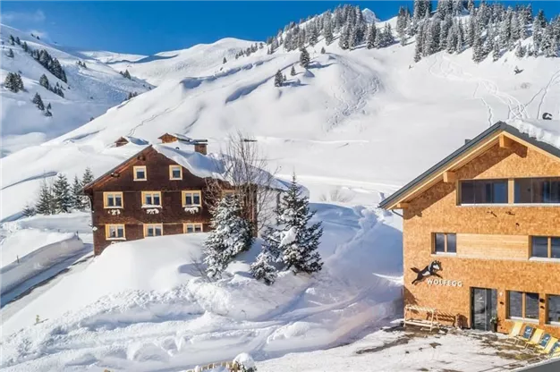 A snowy mountain landscape with two cozy chalets. The trees are covered with snow and the sky is clear and blue.
