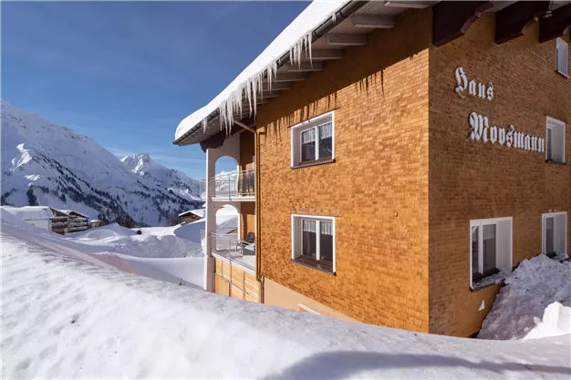 A cozy wooden house in the Alps, surrounded by sparkling snow. In the background, snow-covered mountains can be seen under a blue sky.