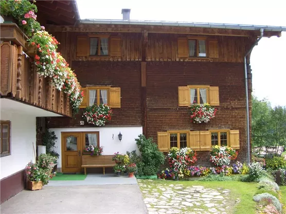 Ein charmantes Holzhaus mit bunten Blumenkästen an den Fenstern. Der Vorbereich ist mit Blumen und einer Bank liebevoll gestaltet.