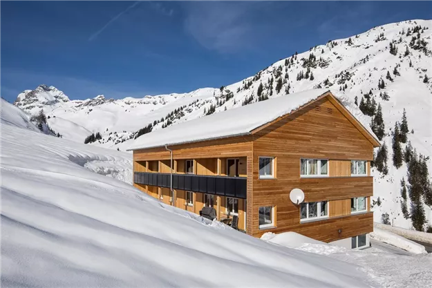 Ein Holzhaus steht inmitten einer verschneiten Landschaft. Im Hintergrund sind schneebedeckte Berge und ein klarer blauer Himmel zu sehen.