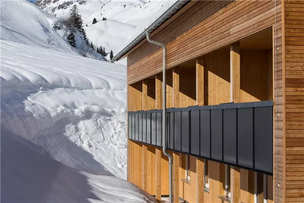 Ein modernes Holzgebäude in einer schneebedeckten Landschaft. Im Hintergrund sind schneebedeckte Berge zu sehen.