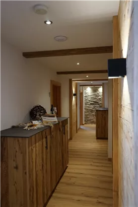 A rustic hallway with bright wooden surfaces and modern lighting. At the end of the hallway, decorative stone walls are visible.