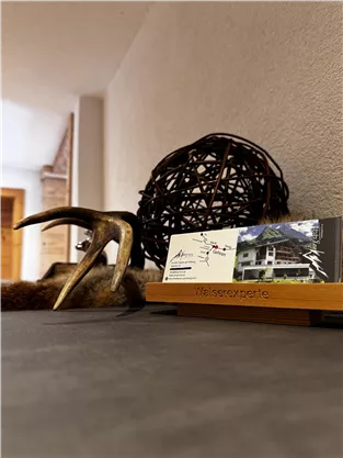 A rustic table with a decorative wooden sculpture and an antler. On it are business cards for an accommodation in the Alps.