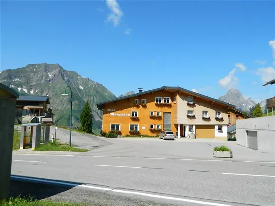 A cozy building in a mountain landscape with a blue sky. In the foreground, there is a road and some cars visible.