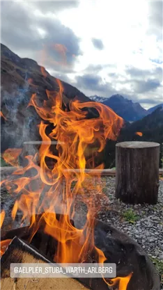A glowing fire burns in the foreground, while the mountains and the cloudy sky are visible in the background. The scene exudes a cozy and nature-friendly atmosphere.