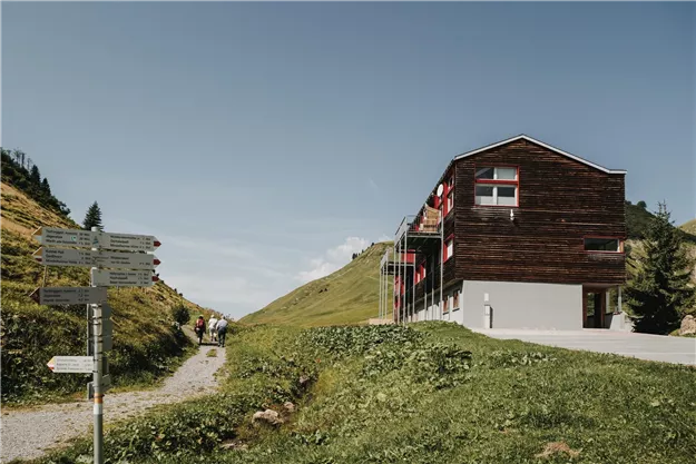 A modern wooden building stands in a hilly landscape with green meadows. Hiking trails and a clear sky enhance the idyllic scenery.