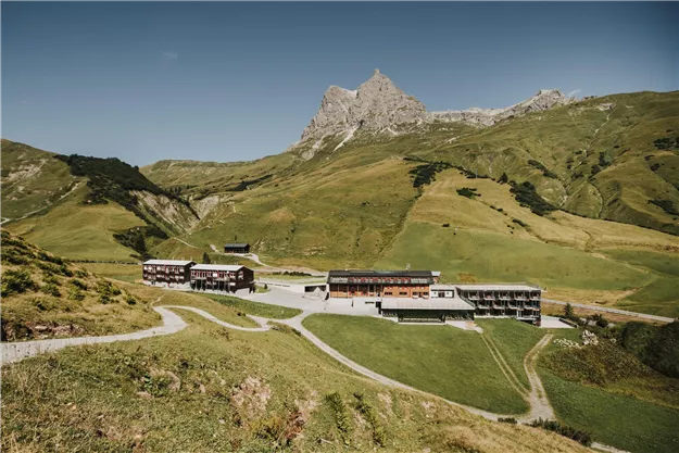 A picturesque mountain landscape with gentle hills and tall mountains in the background. In the foreground are modern buildings that are integrated into the natural environment.