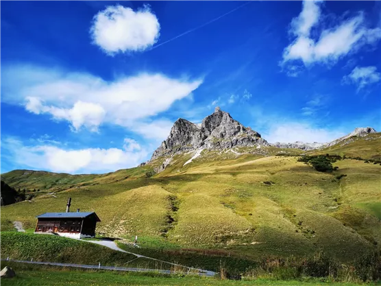 A picturesque mountainous landscape with rugged peaks and a meadow. In the foreground, there is a cozy cabin under a clear blue sky.