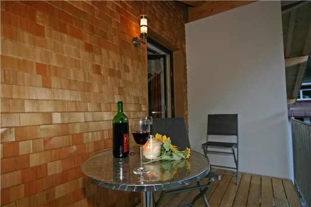 A cozy balcony with a round table, a bottle of wine, and a glass. In the background, wooden paneling and a chair are visible.