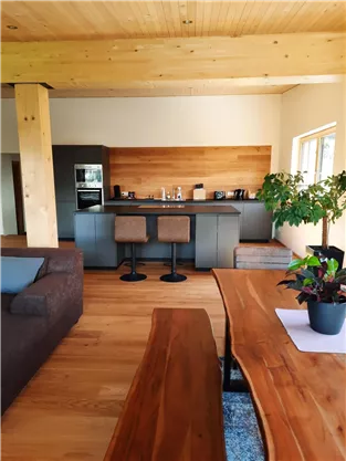 A modern living room with wooden flooring and large windows. The open kitchen has an elegant countertop and is well-equipped.