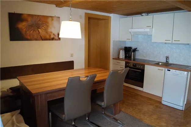 A modern kitchen with a large wooden table and elegant chairs. The walls are bright and adorned with a decorative picture.