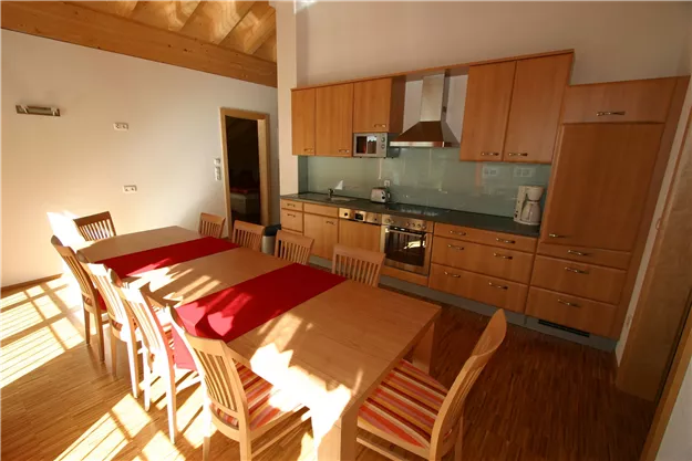 A modern kitchen with wooden cabinets and a large dining table. The room is bright and inviting, with natural light.