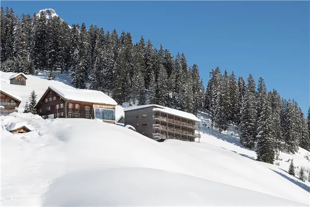 A snowy winter landscape with several huts and tall trees. The clear blue sky provides a beautiful contrast to the white snow.