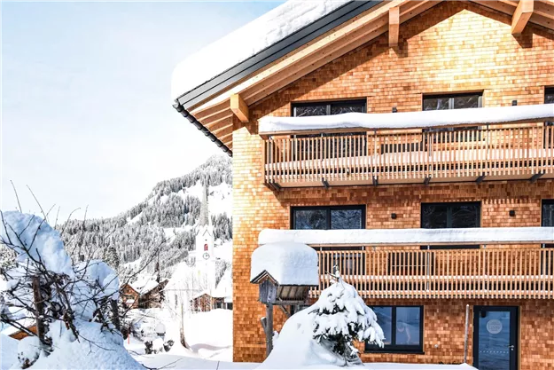 Ein gemütliches Holzhaus inmitten einer schneebedeckten Landschaft. Die Balkone sind mit frischem Schnee bedeckt und bieten einen Blick auf die Berge.