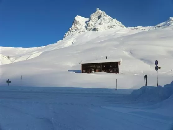 A remote cabin in the midst of a snowy landscape, surrounded by tall mountains. The sky is clear and blue.