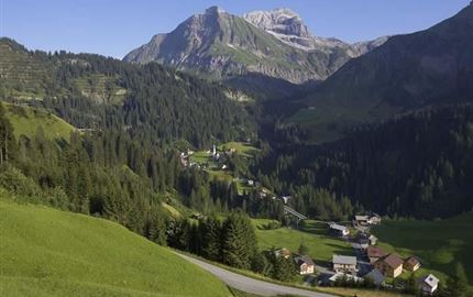 Eine malerische Alpenlandschaft mit grünen Wiesen und hohen Bergen. Im Vordergrund sieht man ein kleines Dorf mit rustikalen Häusern.