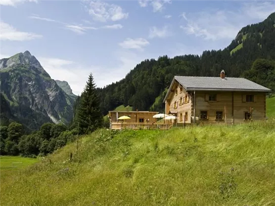 Ein rustikales Holzhaus in einer grünen Landschaft. Im Hintergrund sind majestätische Berge und ein klarer Himmel zu sehen.