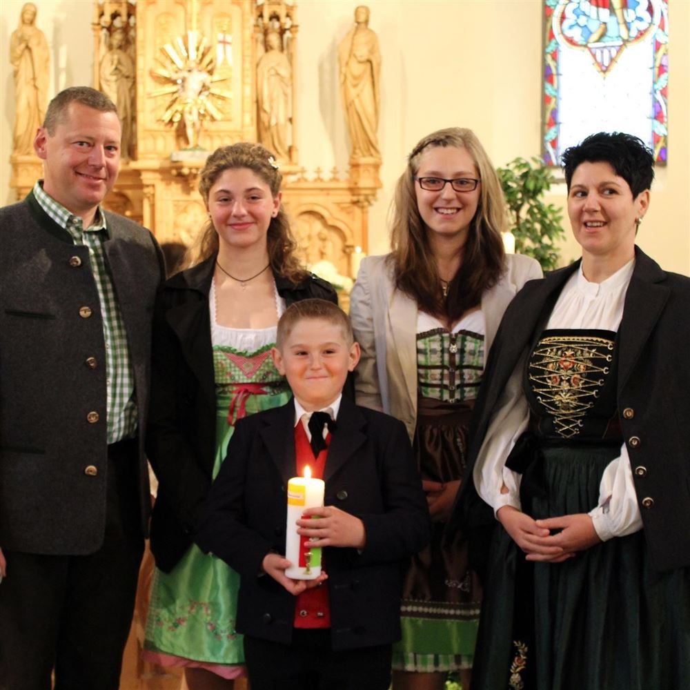 Eine Familie steht in einer Kirche, fröhlich zusammen mit traditioneller Kleidung. Ein Junge hält eine Kerze in der Hand, während die Erwachsenen lächeln.