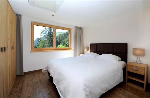 A modern bedroom with a large window that offers a view of green trees. The room is simply furnished with a double bed and a nightstand.