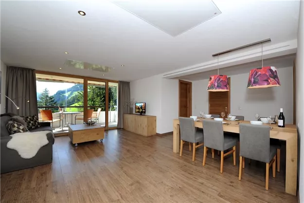A modern living room with a dining table, chairs, and large windows offering a view of the mountains. The decor is bright and friendly, ideal for relaxing stays.