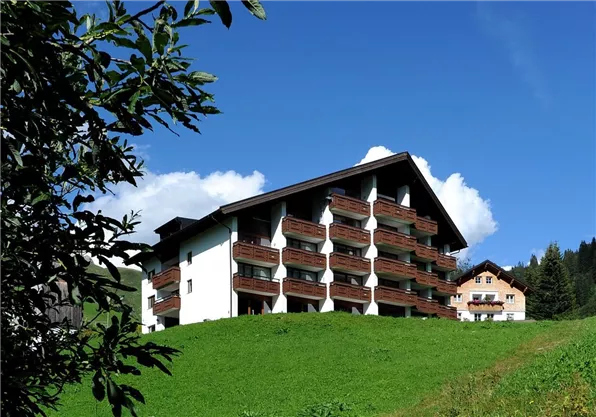 A modern building on a green meadow, surrounded by trees and mountains. The sky is clear and sunny.