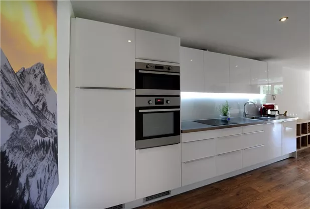 A modern kitchen with white furniture and modern appliances. On the wall, there is a picture of mountains.