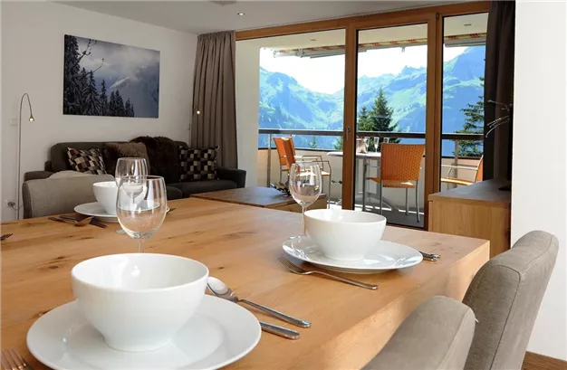 A modern dining room with a large wooden table and elegant dishes. Through the windows, there is a breathtaking view of the mountains.