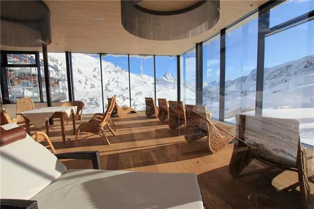 A modern restaurant with large windows and a view of snow-covered mountains. Cozy wooden and wicker furniture creates an inviting atmosphere.