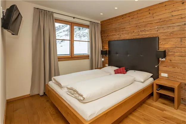 A modern bedroom with wooden paneling and large windows. The double bed is cozy and equipped with white linens.