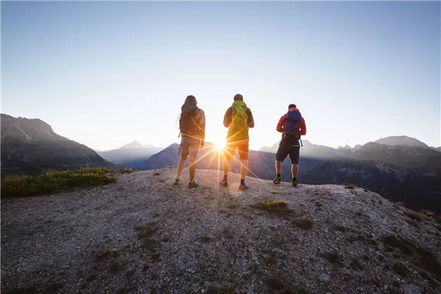 Three hikers are standing on a mountain peak, admiring the sunrise. The landscape is breathtaking with mountains in the background.