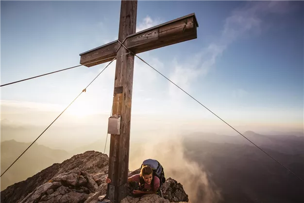 A hiker reaches the summit of a mountain and leans over a large wooden cross. In the background, clouds and mountains are visible.