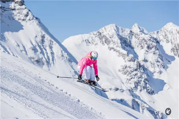 A female skier in pink clothing is skiing down a snowy slope. In the background, impressive snow-covered mountains can be seen.