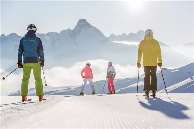 A group of skiers stands on a snow-covered slope, looking at the impressive mountains. The sky is clear and the sun is shining.