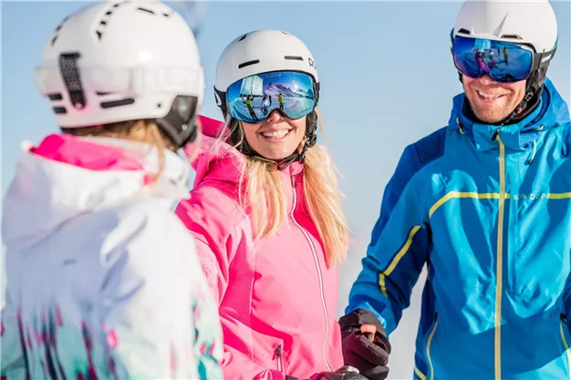 A group of skiers in colorful ski outfits stands in the sun. They laugh and enjoy their time in the mountains.