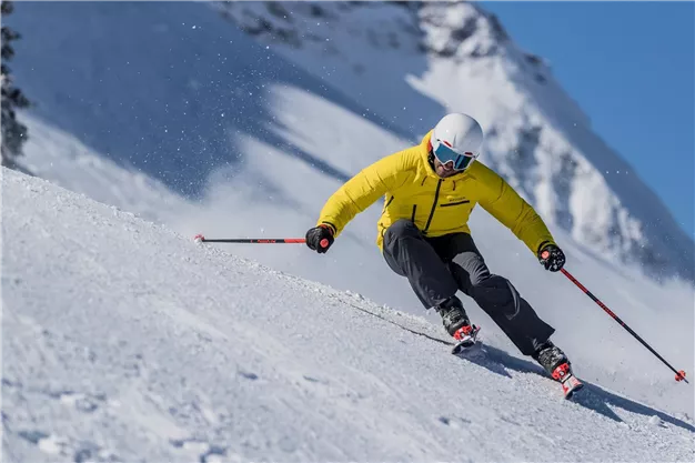 A skier in a yellow suit skis down the snow-covered slope. In the background, snow-covered mountains and a blue sky can be seen.