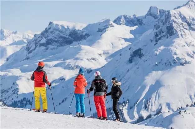 A group of skiers stands on a snowy mountain peak. In the background, majestic mountain peaks and a blue sky can be seen.