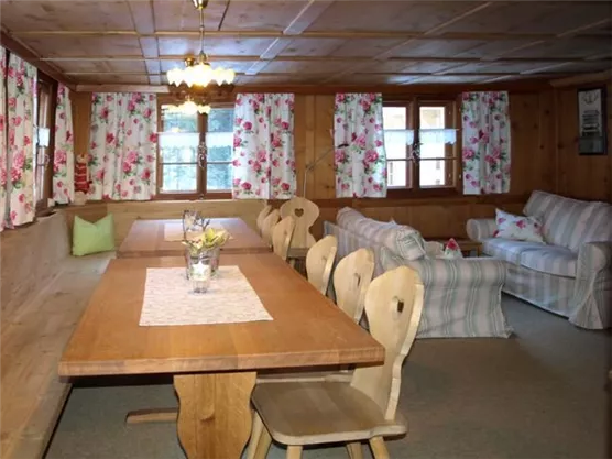 Ein gemütlicher Wohnraum mit Holzverkleidung und blumigen Vorhängen. Ein großer Esstisch und eine bequeme Couch laden zum Entspannen ein.