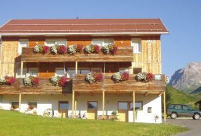 Ein Holzgebäude mit mehreren Balkonen, geschmückt mit bunten Blumen. Im Hintergrund sind grüne Wiesen und ein Berg zu sehen.
