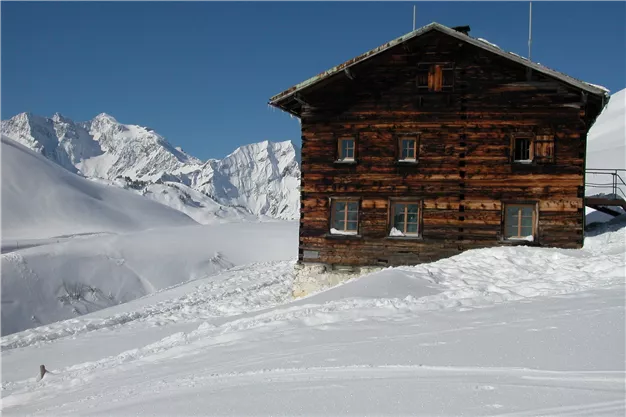 An old wooden house stands in the snowy landscape of the mountains. The clear blue sky highlights the majestic peaks.